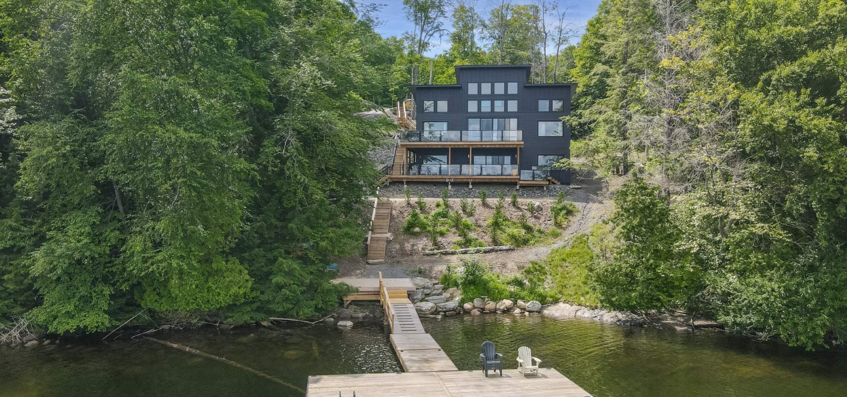 Cottage surrounded by trees with 2 levels of decks overlooking the lake. Stairs leading to a long dock with 2 chairs on it.