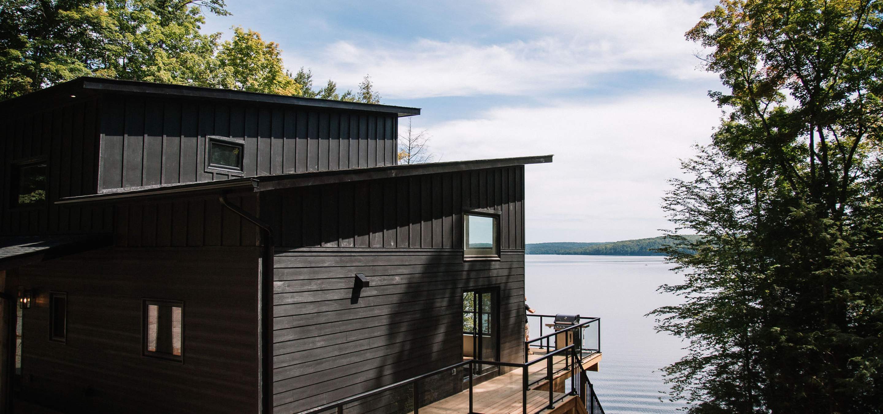 cottage with decking around it, facing the lake with trees around it. 