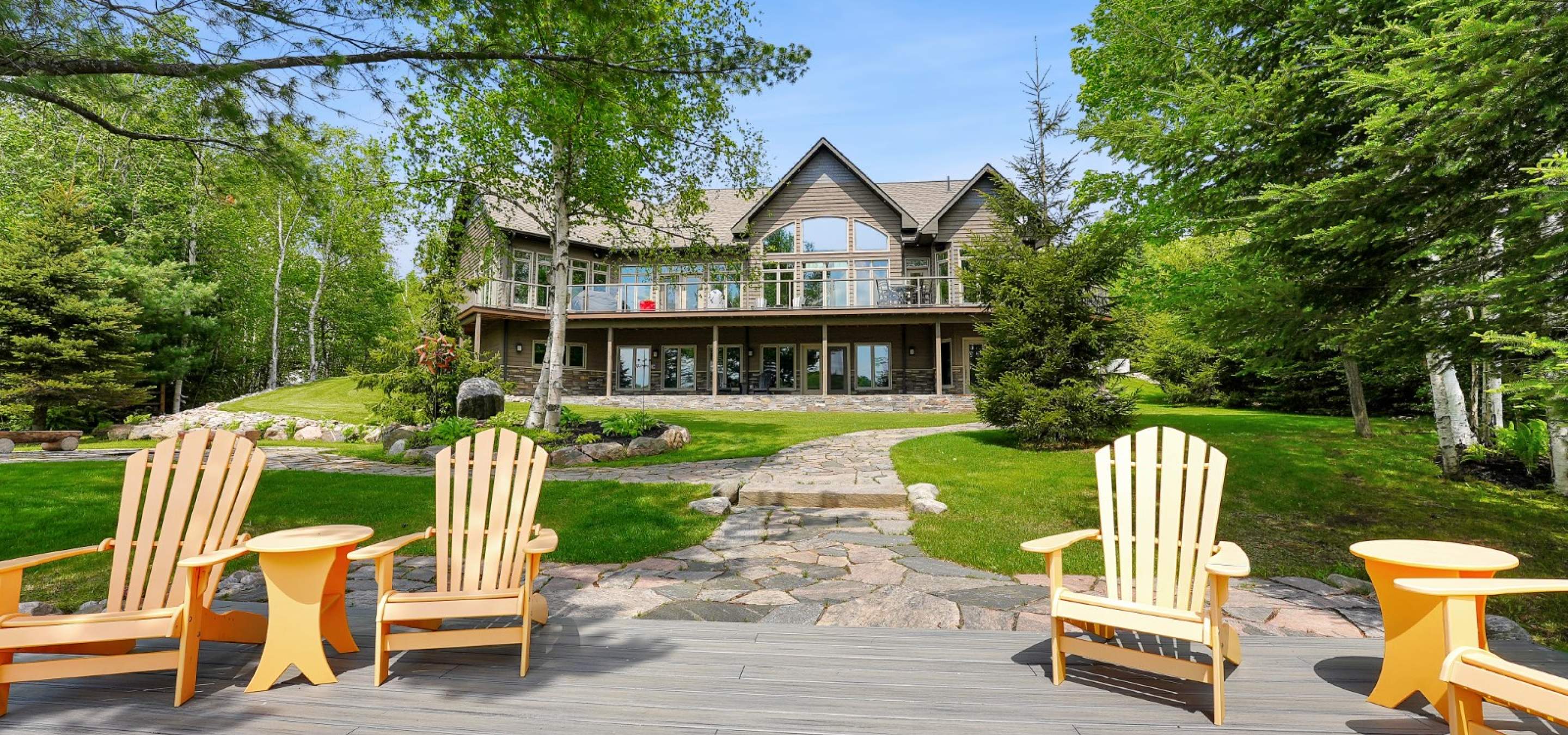Cottage with lots of windows, green grass and trees Stone path leading from cottage to dock with 4 yellow muskoka chairs