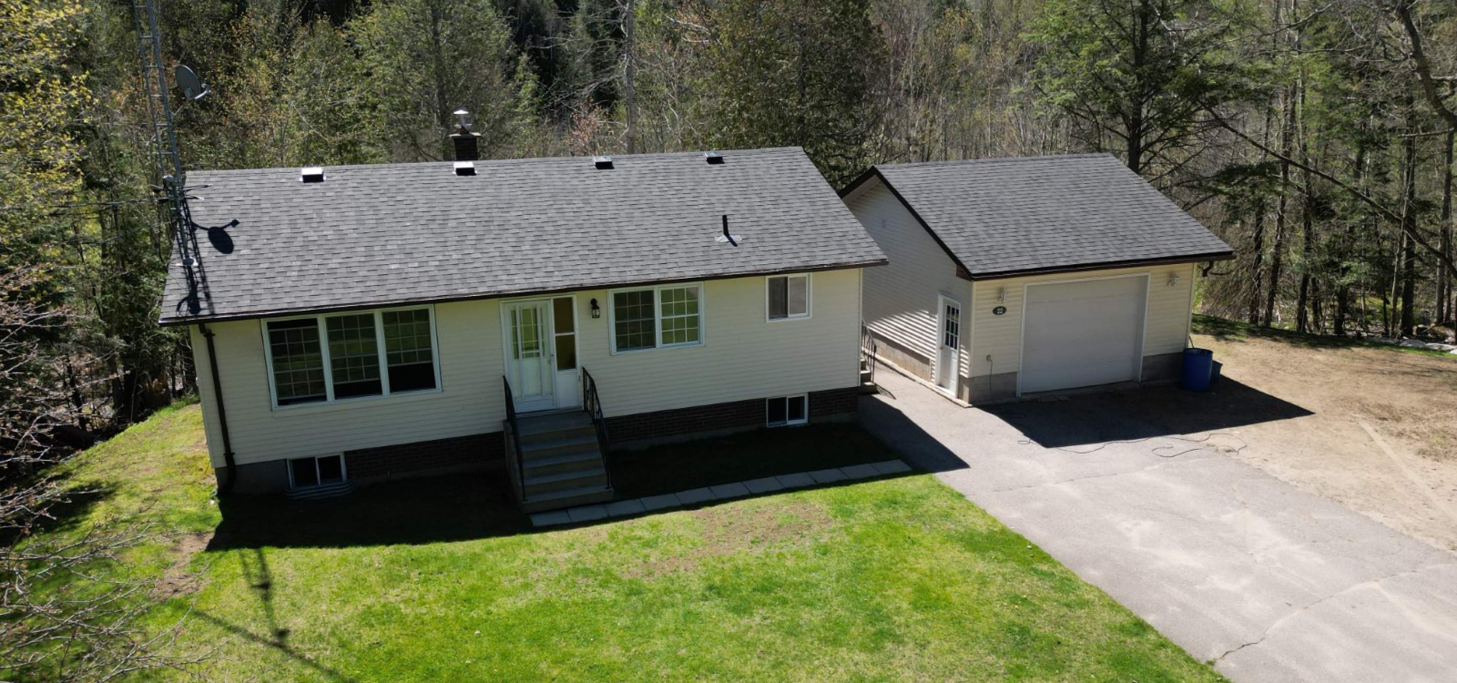 Bungalow with garage on the right, paved driveway, green lawn and forest perimeter.