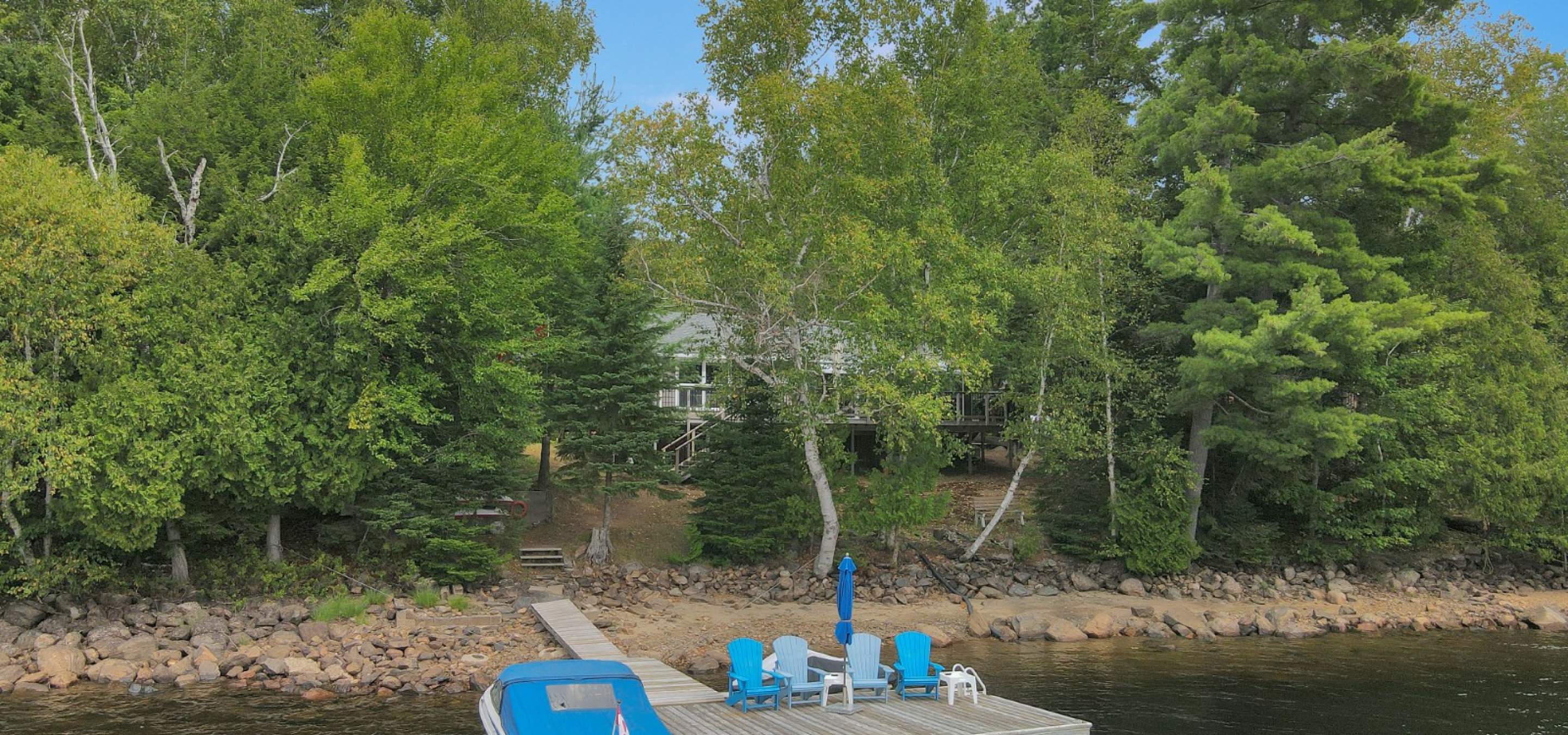 Cottage hidden behind trees with sandy beach front and dock with 4 blue chairs and boat with blue cover.