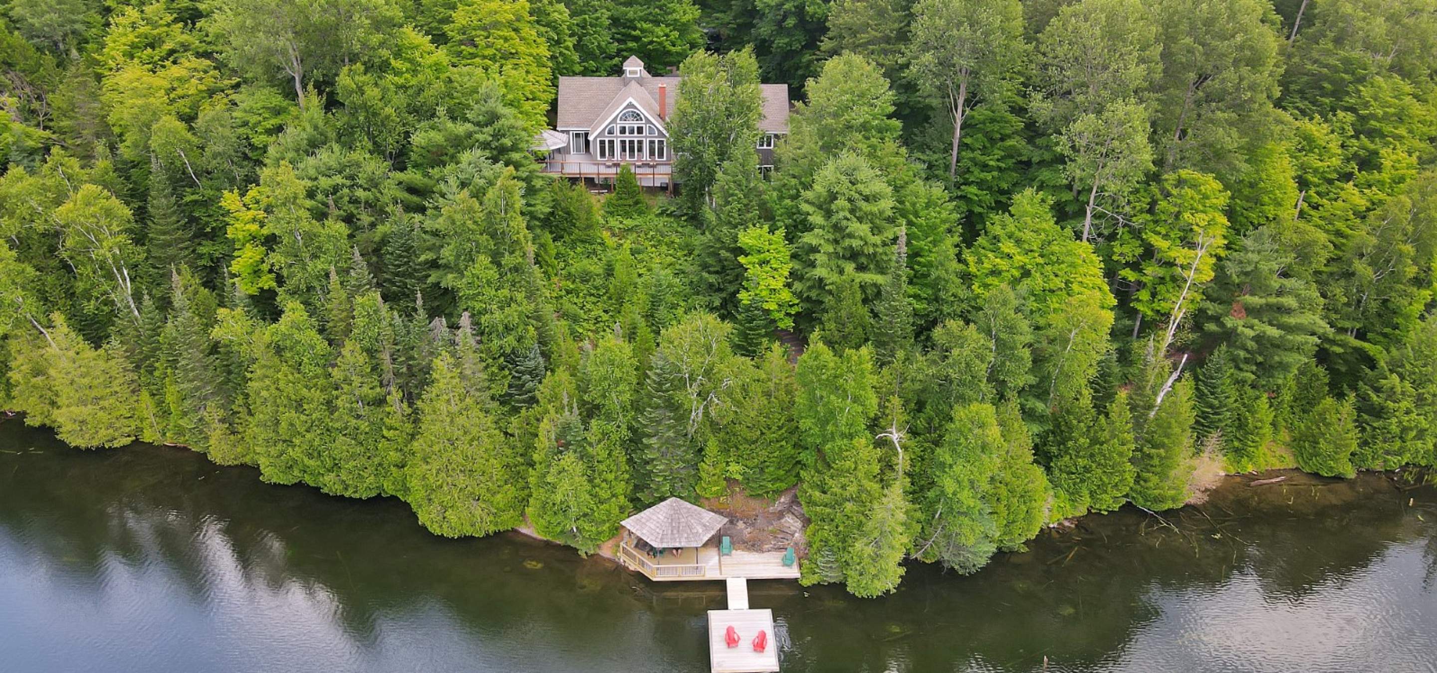 Home atop a forested hill with a gazebo deck and dock with 2 red muskoka chairs at the lake below