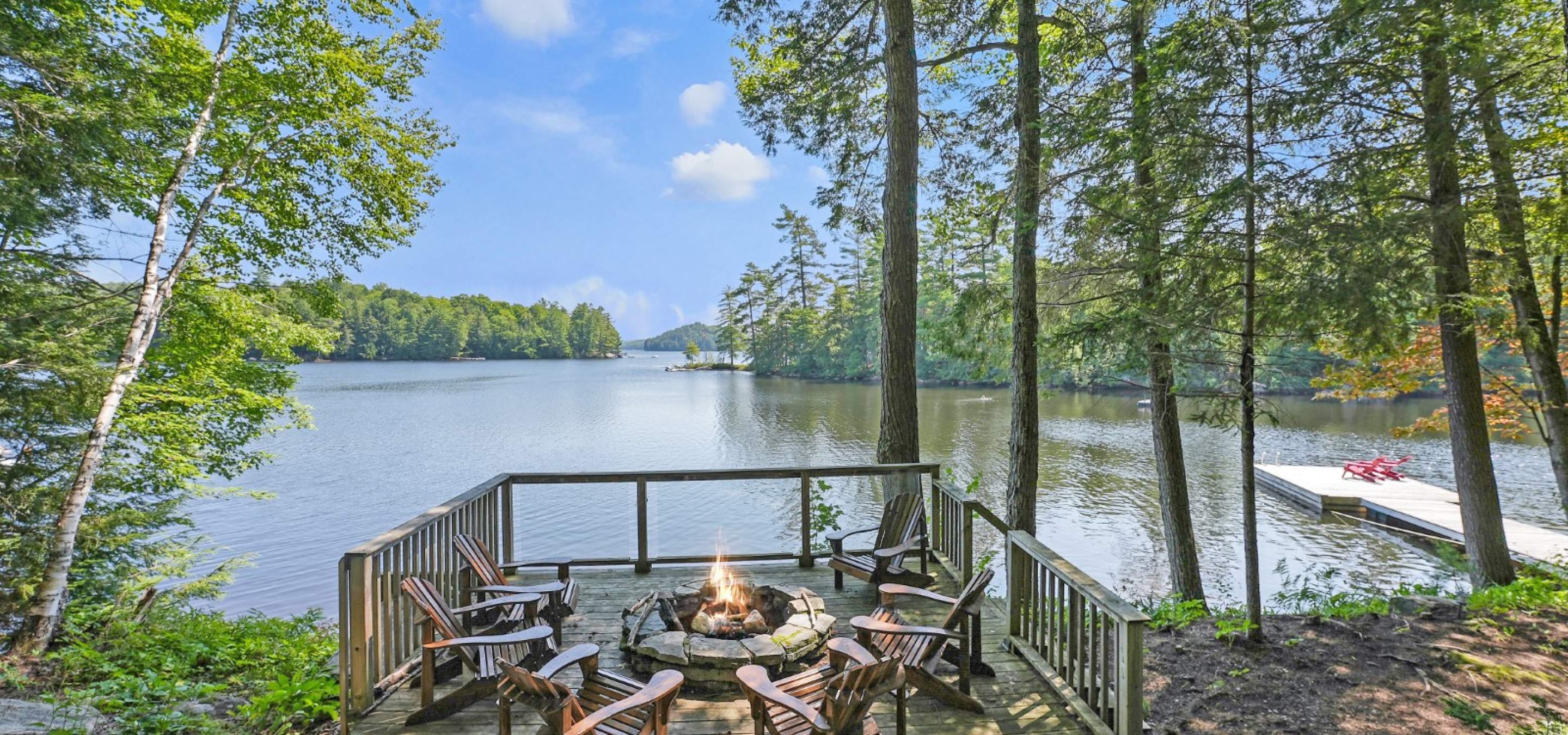Fire pit with 6 chairs around it and a glass railing overloooking a lake. Trees surround and a dock in the water with red chairs.