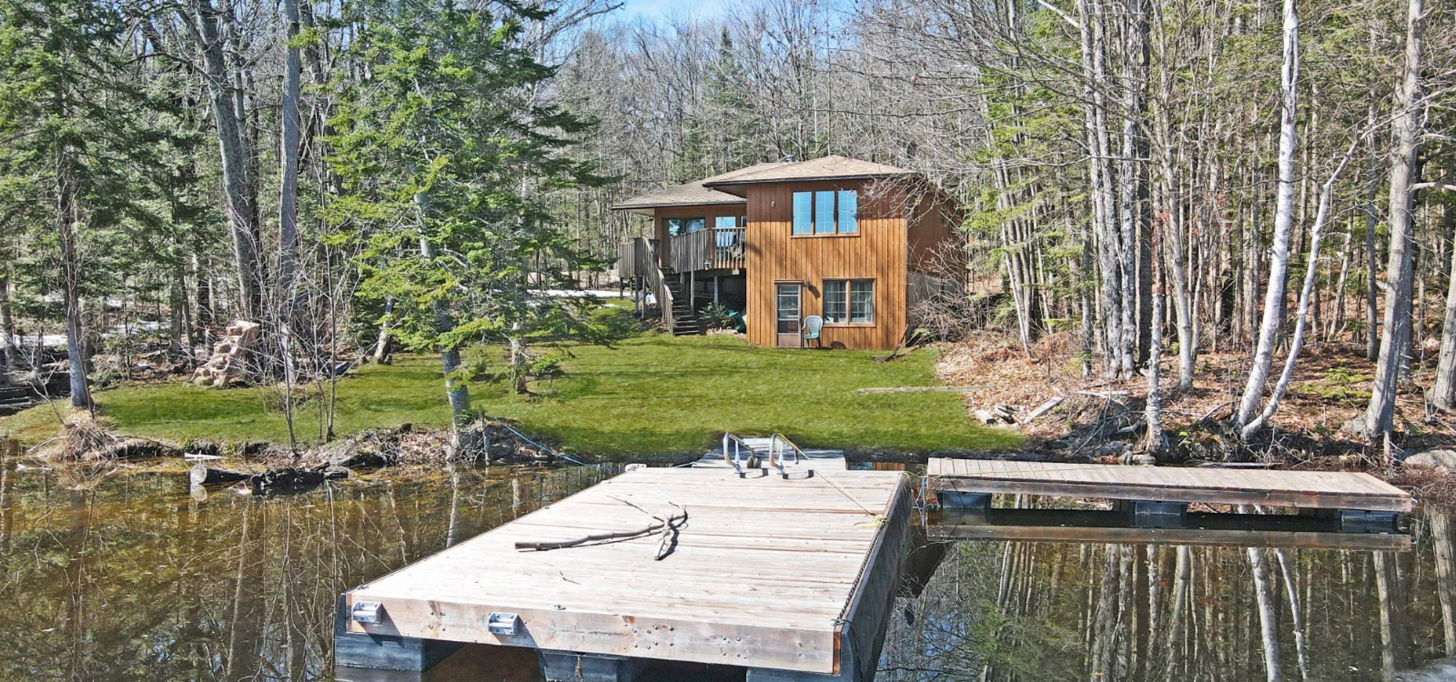 Wood cottage with grass, surrounded by trees with a dock into the lake.