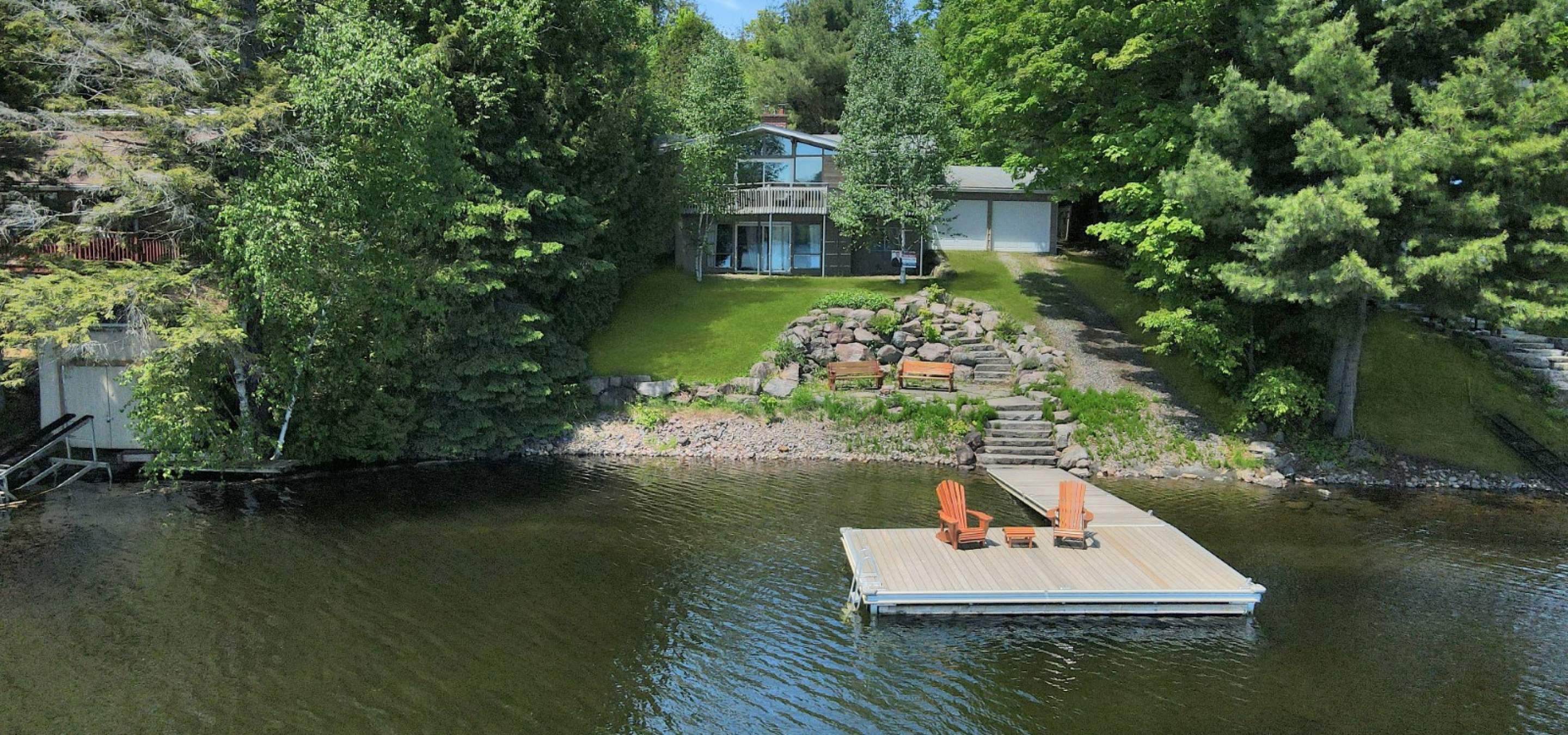 cottage with 2 tier deck and double car garage. Trees on either side with grass in the middle. Dock with 2 orange muskoka chairs.