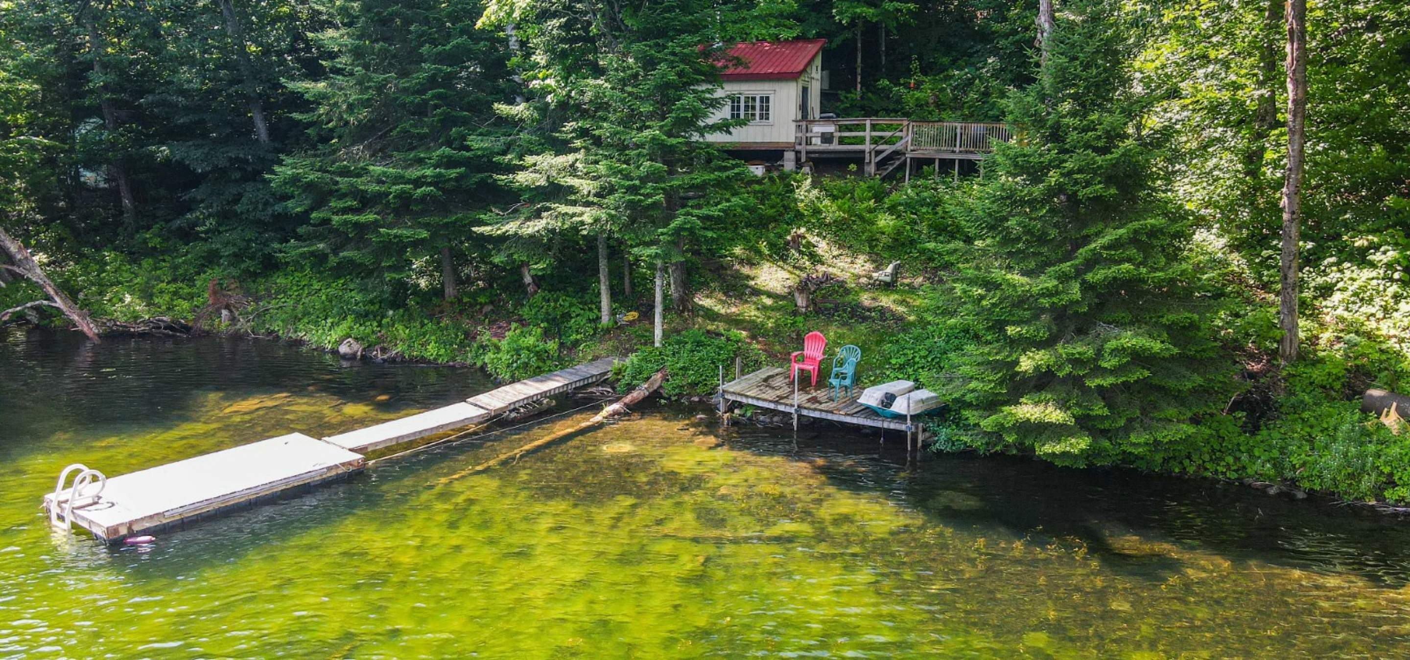 Yellow cabin with red roof hidden behind trees, grass hill leading to a long dock out into the water, and a red & green muskoka chair.