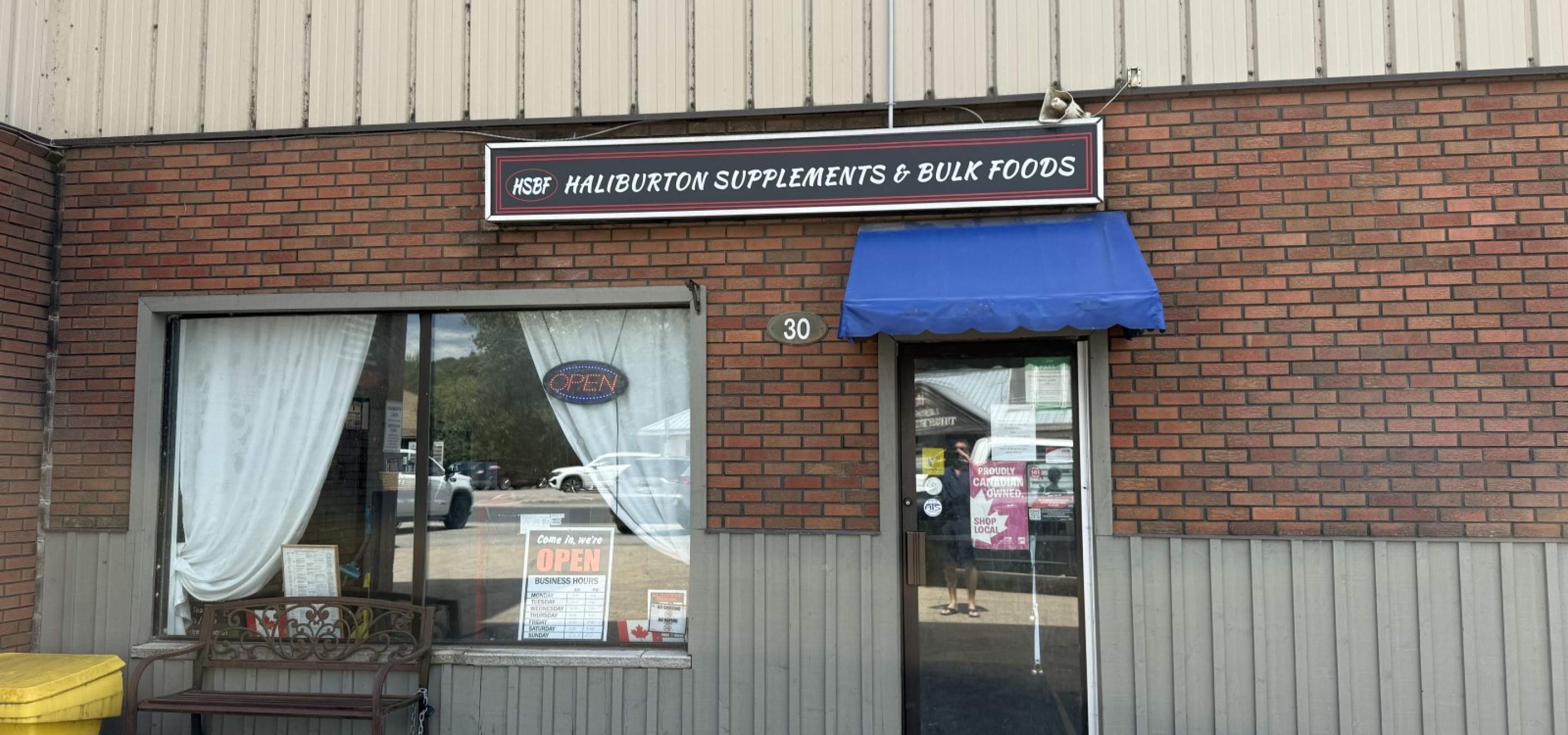 Brick store front with large window, glass door with blue awning. Business sign reading Haliburton Supplements and Bulk Foods