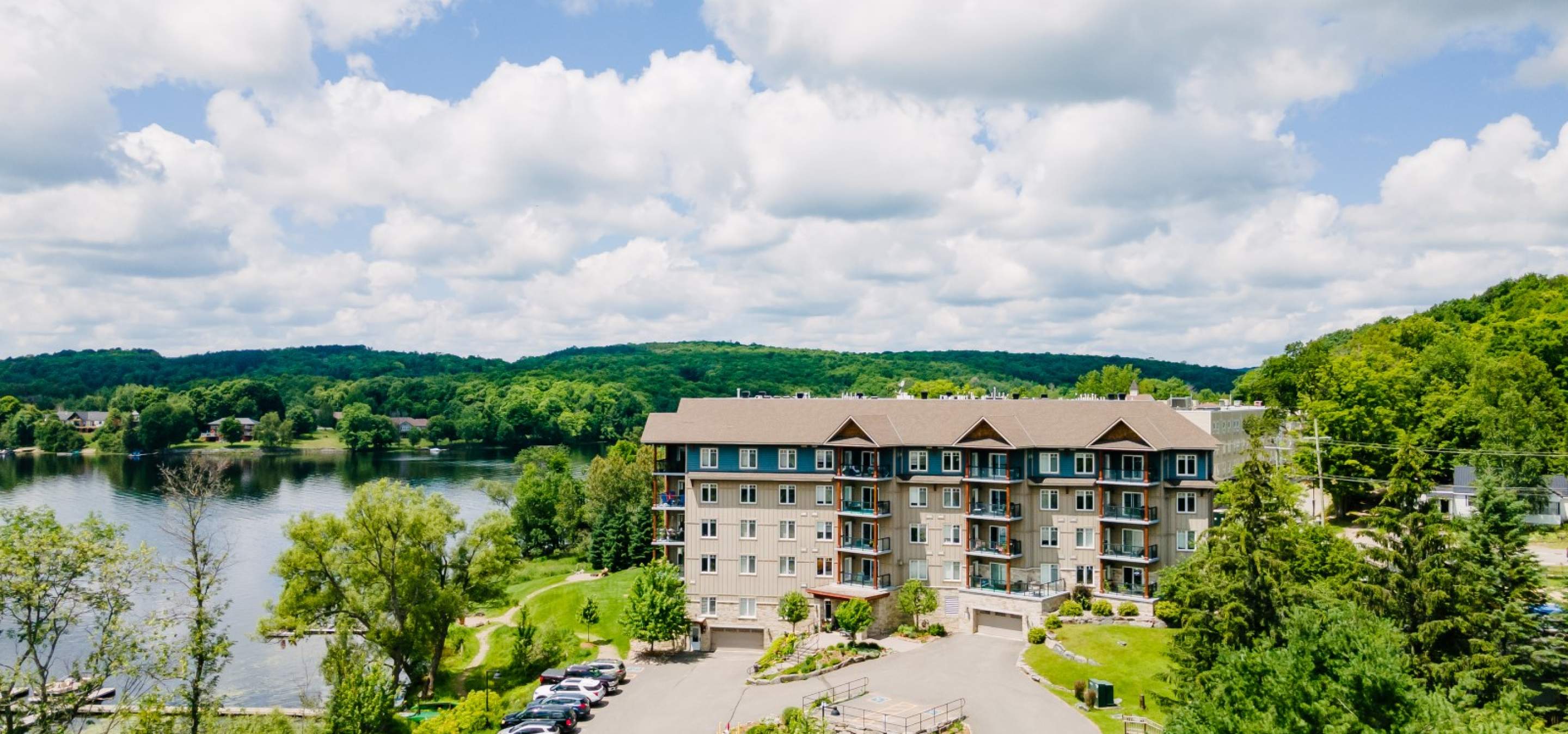 condo building with a parking lot, surrounded by trees and green grass, located beside a lake. 
