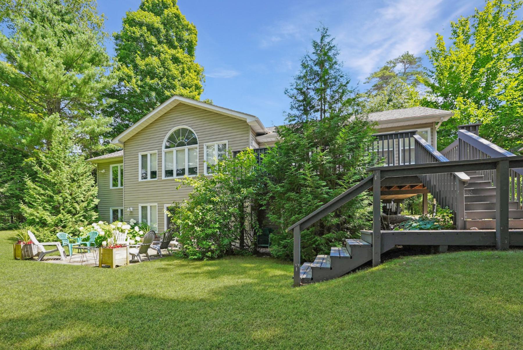 2 level home with deck and stairs on the right to green manicured lawn and patio with chairs at the front of the home