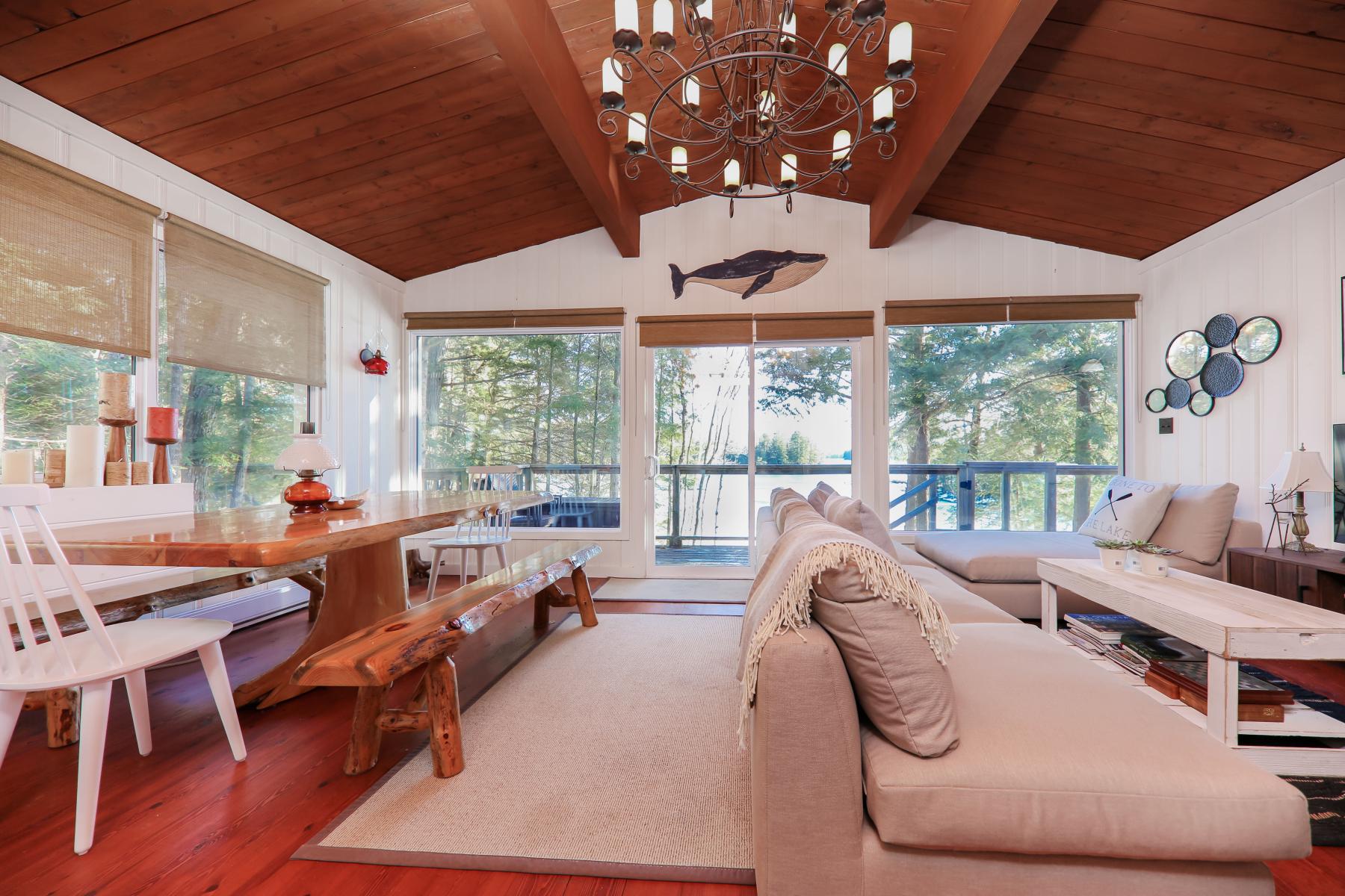 interior of a cottage with wood floors, wood ceiling, live edge dinning table with bench and 5 big windows looking at trees. 