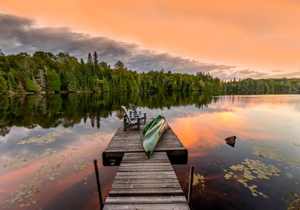 Muskoka chairs and a canoe sit on a dock overlooking a lake and treed shoreline at sunset. 