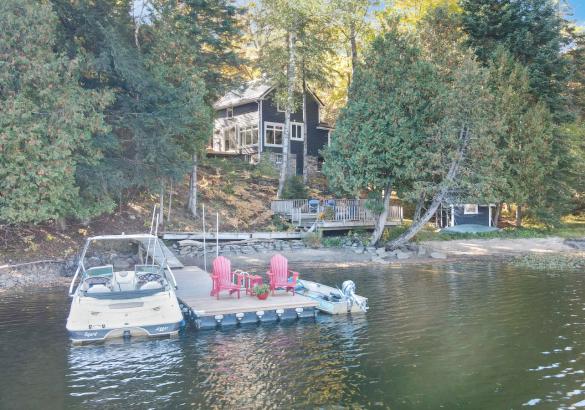 look at a cottage from the lake with 2 boats and 2 red chairs on the dock.