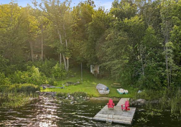 View from Salerno Lake toward the waterfront cottage, with a dock, two red chairs, boat and grassy shoreline.