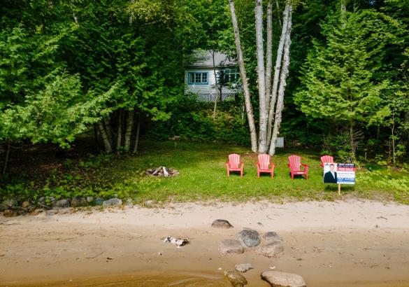 white cottage hidden by green trees. Grass lawn with 4 red muskoka chairs and a sandy beach front. 