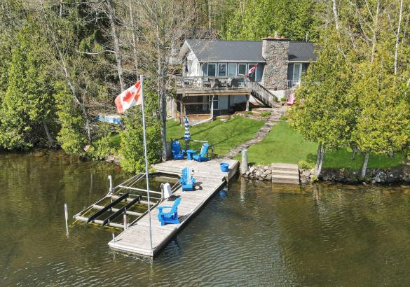 White cottage with a stone chimney with wrap around deck. Surrounded by trees with a dock in front featuring 4 blue chairs. 
