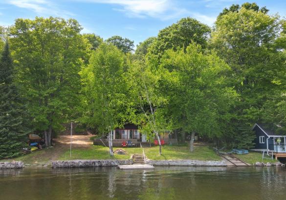 looking at a cottage from the water, large trees, stone wall, and boat launch