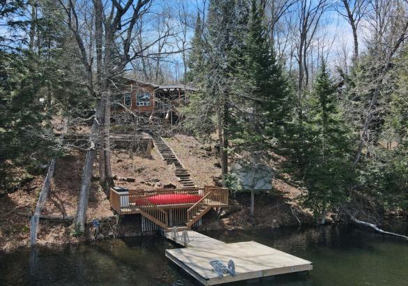 Cottage on a hill behind trees, stairs down to the dock in the water, and a red canoe.
