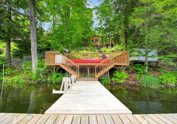 Cottage on a hill behind trees, stairs down to the dock in the water, and a red canoe.