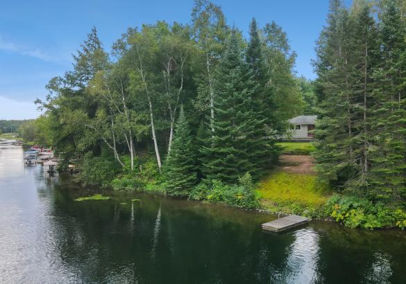 view of a grey house from the river with a dock, sandy area and trees