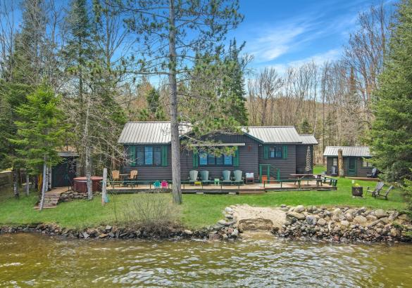 Grey cottage behind some trees with a the lake infront and green grass.