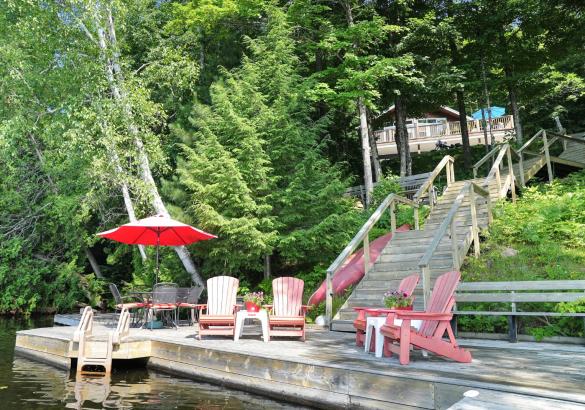 Dock with muskoka chairs with a red umbrella. Stairs leading up to the lakeside home amoungst trees