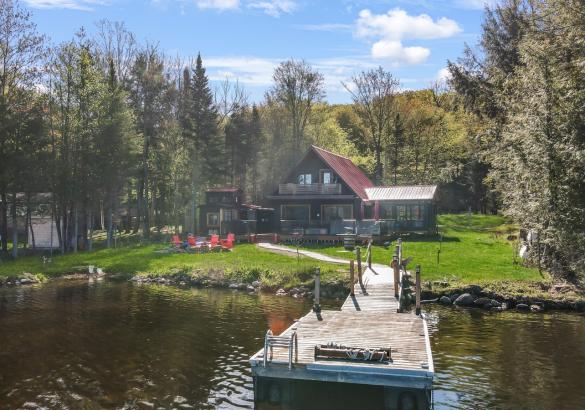 a frame cottage with green grass, red chairs around the fire, sand beach and dock into lake.