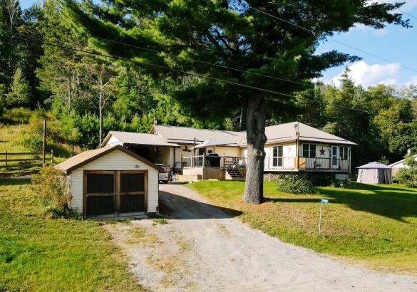 Home surrounded by trees with a grass front lawn and large deck. Gravel laneway leading to home and garage.