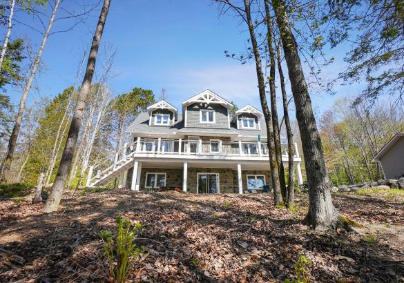 Gaze up at a blue sky and majestic grey home with white trim, deck across the front and 2 dormers on the upper level with tall trees around