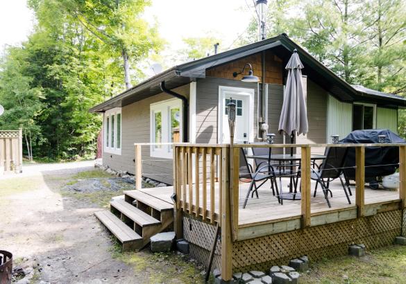 Wood deck with patio set and barbecue, tidy cottage with trees in the background.