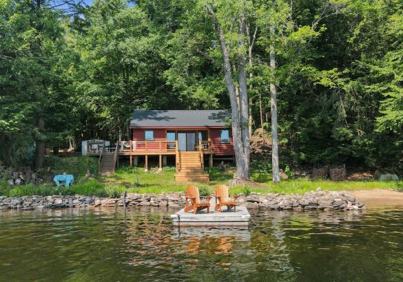 Red cottage in the trees with stairs leading down to grass. 2 muskoka chairs on dock in front.