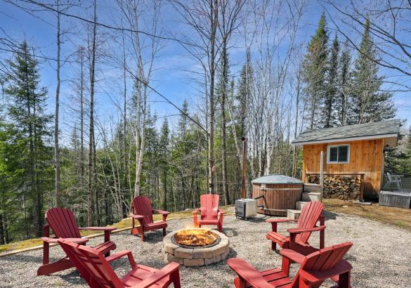 6 red muskoka chairs around a campfire and a bunkie in the background