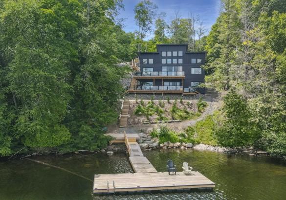 Cottage surrounded by trees with 2 levels of decks overlooking the lake. Stairs leading to a long dock with 2 chairs on it.