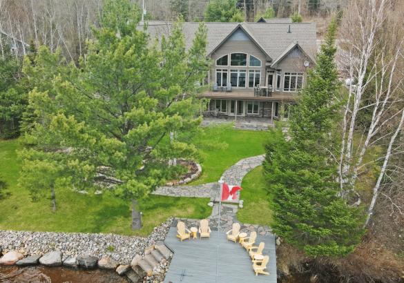 Cottage with lots of windows, green grass and trees Stone path leading from cottage to dock with 6 yellow muskoka chairs and canadian flag. 