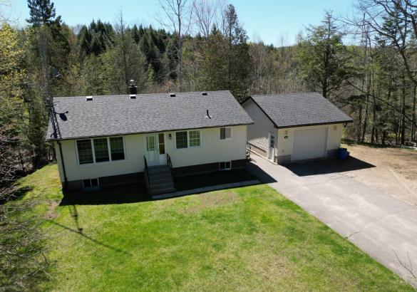 Bungalow with garage on the right, paved driveway, green lawn and forest perimeter.