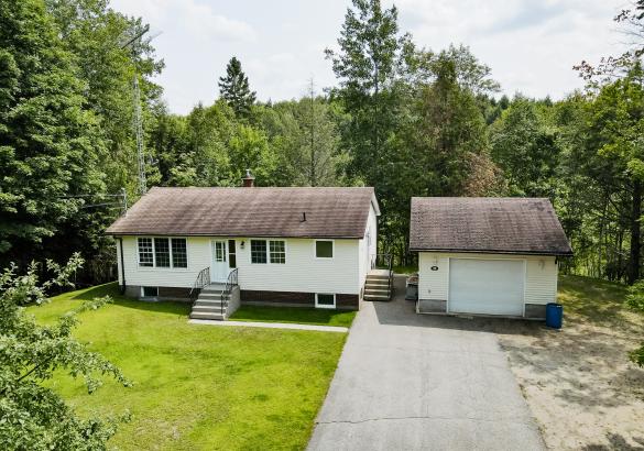Bungalow with garage on the right, paved driveway, green lawn and forest perimeter.