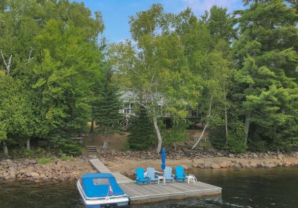 Cottage hidden behind trees with sandy beach front and dock with 4 blue chairs and boat with blue cover.