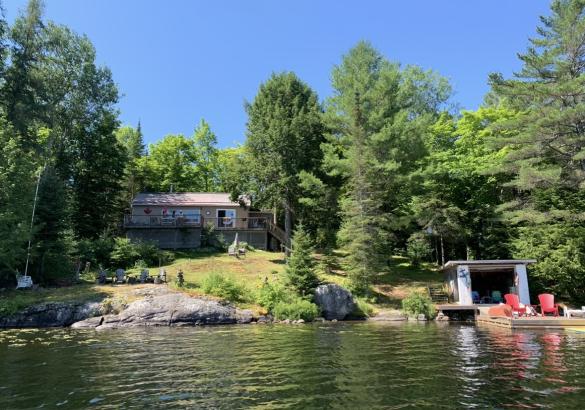 Cottage from the lake with trees, a boat house and 2 red chairs