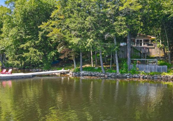 lakefront cottage surrounded by trees with a dock out into the lake. 2 red muskoka chairs sitting on the dock.