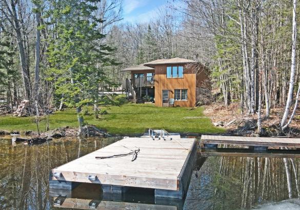 Wood cottage with grass, surrounded by trees with a dock into the lake.
