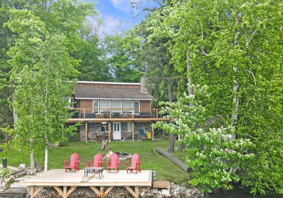 Brown cottage with 2 decks, 4 red muskoka chairs, and large trees.