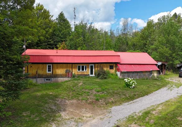 Wood building with red metal roof, gravel laneway, green grass, surrounded by trees.